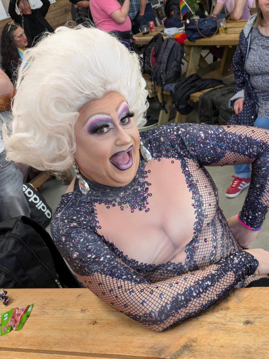 The image shows a Drag Queen wearing a large wig and smiling at the camera