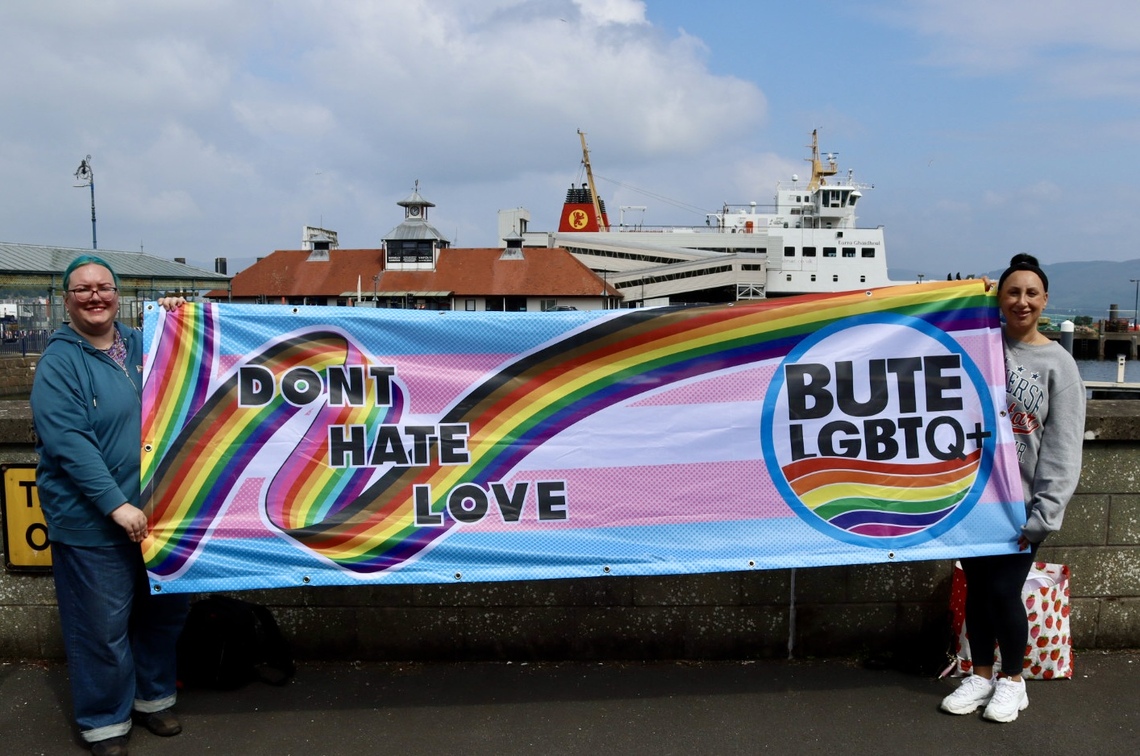 image shows committee members holding a rainbow banner with the ButeLBTQ+ logo and thge words Don't Hate Love