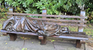 A metal statue of a homeless person wrapped in a blanket lying on a wooden bench