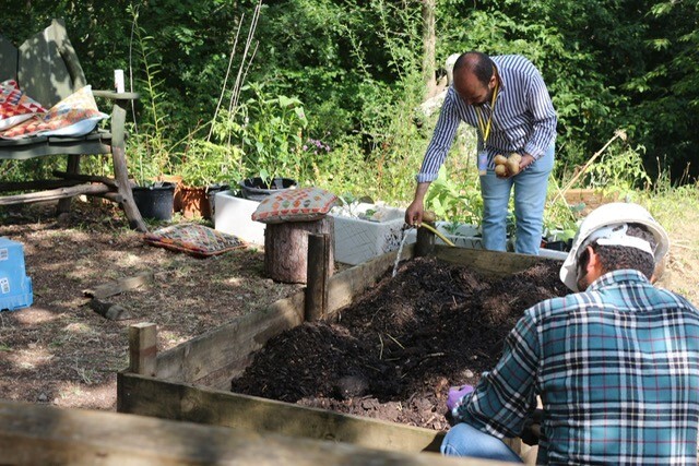 Two men prepare some soil in a raised planter for planting. In the background we see natural wood seating and colourful cushions to sit on