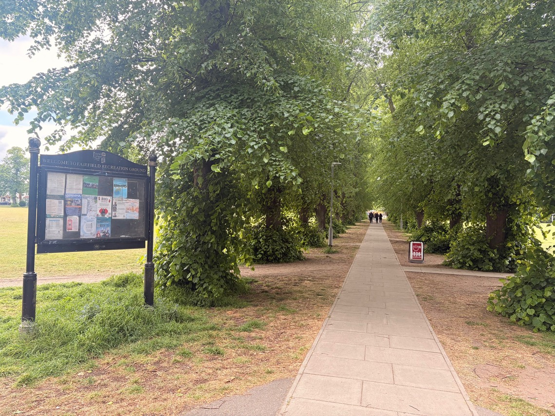 A community noticeboard is in the foreground next to the path cutting through the centre of the park
