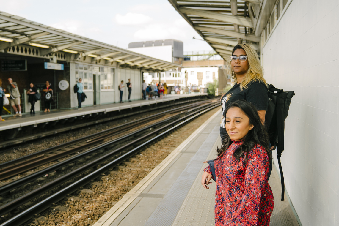 Two women stand on a train station platform looking off camera for the train. By The Unmistakeables