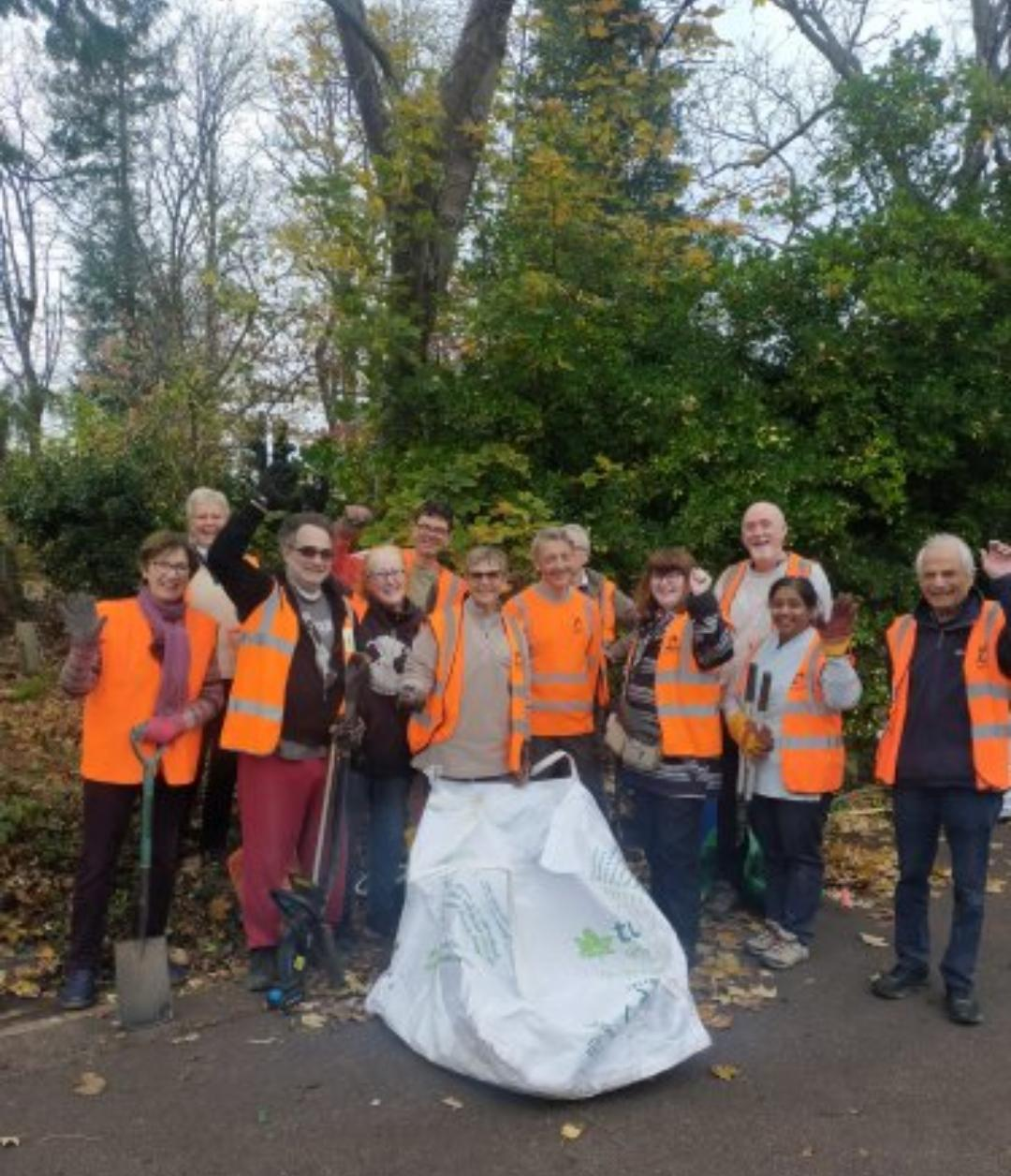 A group of volunteers in high-viz jackets with gardening tools smile at the camera.