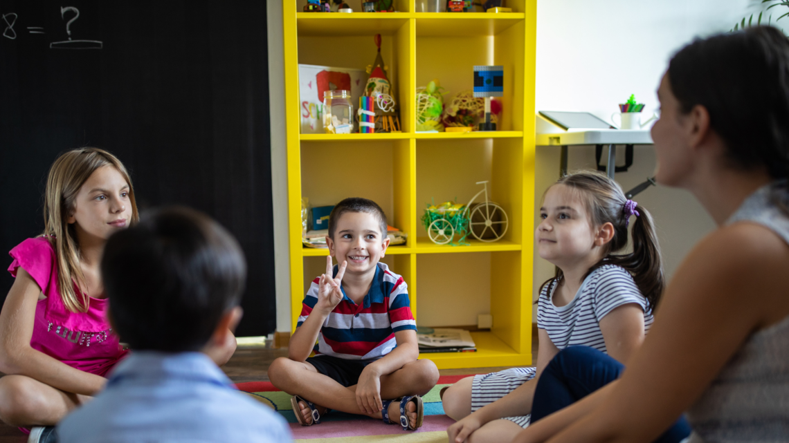 Children sit in a circle on the floor listening to a woman talk