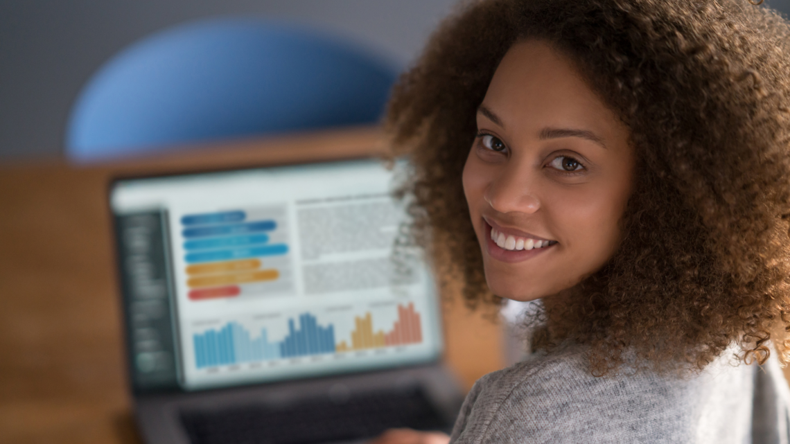 Woman turns face to camera with data on laptop screen in background