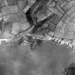 White breaking waves mark Wembury beach. To the east, the dark triangle protruding into the sea is Blackstone Reef, immediately below St Werburgh's Church. The line of development heading north east from the beach is Church Road, with its straggle of ribbon development.  On the cliffs just east of Blackstone Reef a cluster of sheds and old buses overlooks the cliff. Today this site is Cliff Road.