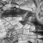 The eastern end of Knighton can be seen in the south west. Traine Road and Wembury Road curve northwards and slightly westwards from Knighton to converge at Hollacombe. Traine Farm can be seen between these two roads. Hollacombe Brake,  the wood running east-west, leads into Wembury Wood on the edge of the sinuous Yealm Estuary. It is low tide - the river is a trickle winding through the mudflats.  